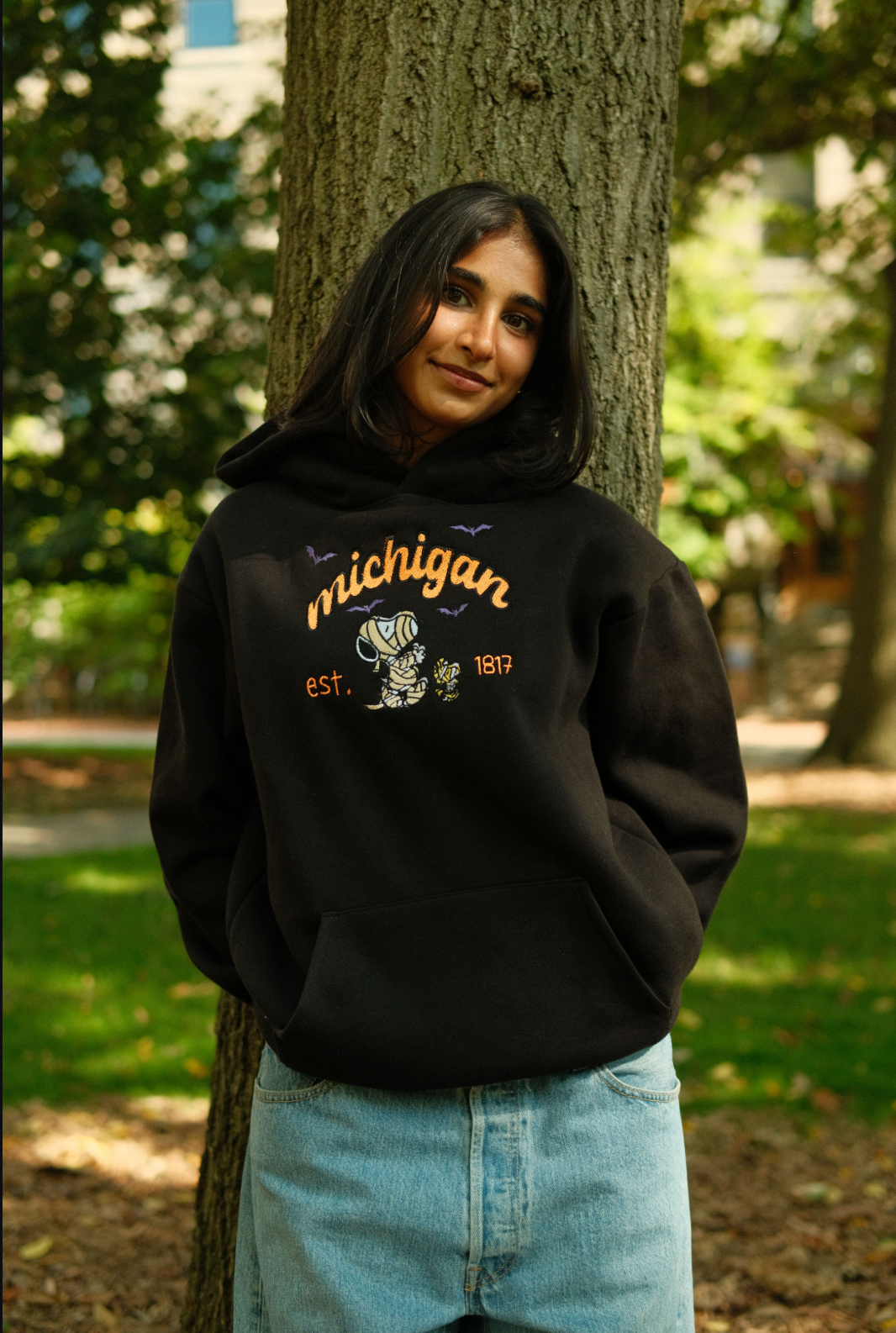 Person wearing a black MITTN hoodie with 'Michigan' and Snoopy design, standing outdoors near trees.